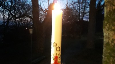 Eine hohe brennende Kerze mit rotem Kreuz und der Aufschrift „202“ steht in einer Außenumgebung bei Dämmerung.