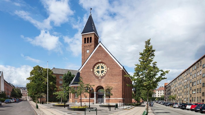 Murstenskirke med højt spir og ur, omgivet af træer og bygninger.