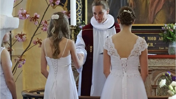 To børn i hvide klæder taler med præst i kirke.