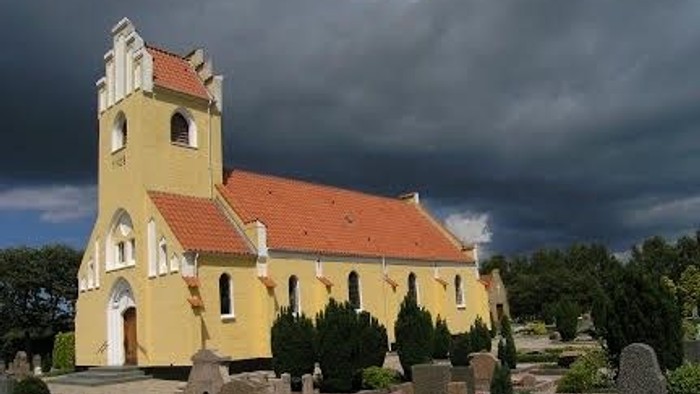 En gul kirke med tårn og rødt tag, omgivet af en gravplads med gravstene og planter.