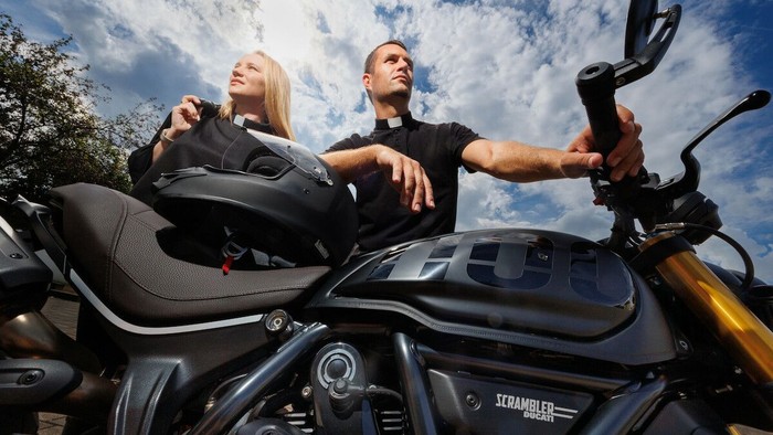 Zwei erwachsene Personen stehen neben einem Motorrad im Freien und blicken in die Ferne. Beide tragen schwarze Oberteile mit sichtbarem Priesterkragen, was auf einen kirchlichen Kontext hinweist. Im Vordergrund dominiert das Motorrad: Der Tank, der Lenker
