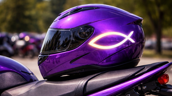 Ein glänzender Motorradhelm in kräftigem Violett liegt auf dem Sitz eines Motorrads. Auf der Seite des Helms ist ein leuchtendes Fischsymbol angebracht, das sich hell vom dunklen Lack abhebt. Das Visier ist geschlossen und spiegelt die Umgebung leicht wid