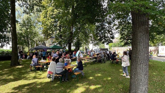 Draußen versammelte Menschen, die an Picknicktischen unter schattigen Bäumen in einer Parkanlage sitzen.