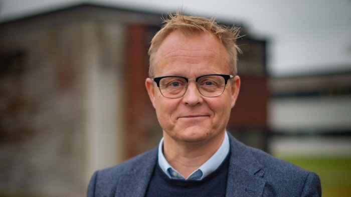 En mand i en mørk jakkesæt og briller står selvsikkert udendørs nær en bygning.