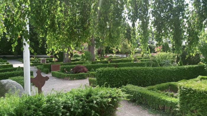 En smuk have med høje buske og træer, en gravsten og en smedestøbe.