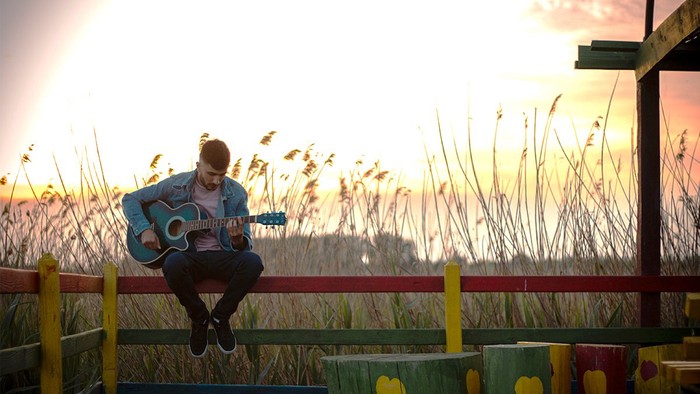 Mann mit Gitarre sitzt auf Zaun vor Abendsonne.