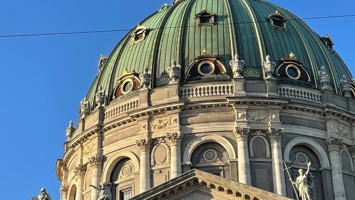 Stor kuppelkirke med latinsk indskrift og klassiske statuer under klar blå himmel.