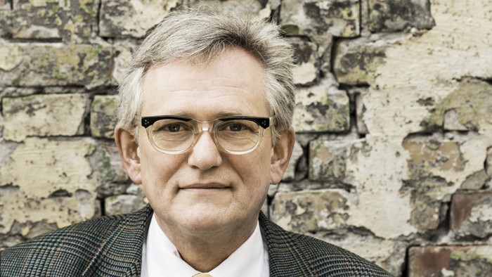 En mand med briller og jakkesæt med et gult slips står op ad en tekstureret stenmur.