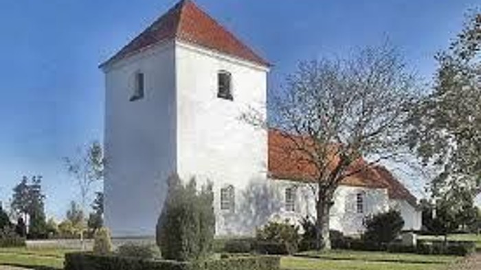 En idyllisk hvid kirke med et rødt tegltårn står midt mellem veltrimmede hække og nøgne vintertræer.