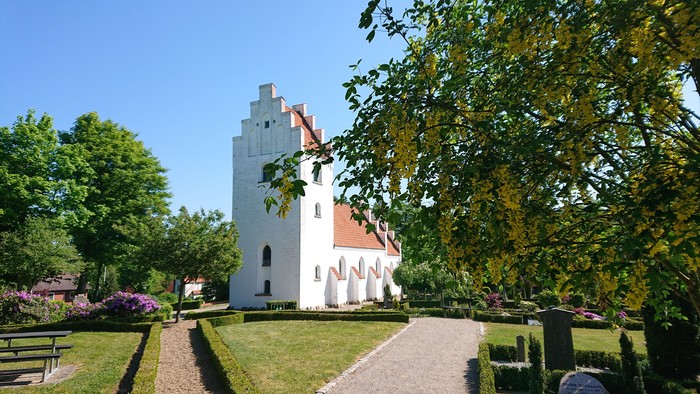 Et hvidt historisk hus med røde tag i en træket garden med bænke og blomster.