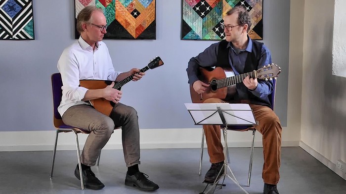 To musikere spiller på strengeinstrumenter, mens de sidder, akkompagneret af noder på et nodestativ.