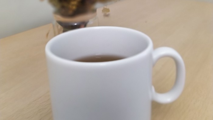 Eine weiße Tasse Tee auf einem Holztisch mit einer bunten Deko und getrockneten Blumen im Hintergrund.