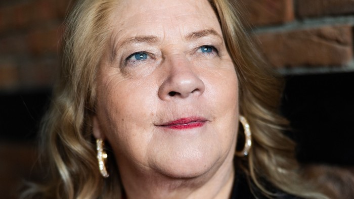 Eine Person mit blondem Haar in einer schwarzen Jacke über einem roten Oberteil vor einer Backsteinmauer.