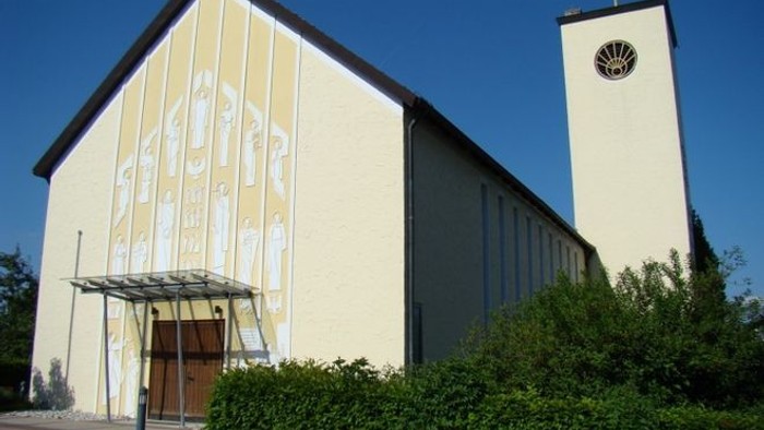 Kleine Kirche mit Glockenturm und Uhr, umgeben von Grün.