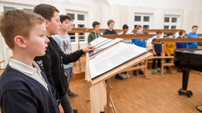Junge Schüler in einem Musikraum proben mit Notenblättern, während ein Dirigent sie anleitet.