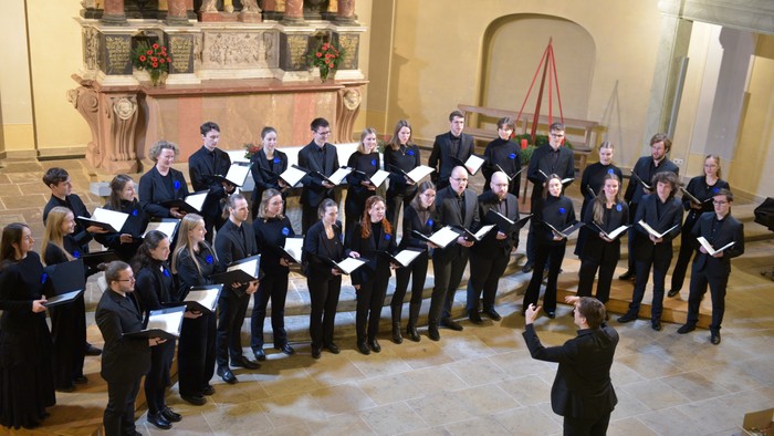 Ein Chor tritt in einer prächtigen Kirche auf, geleitet von einem Dirigenten mit Notenblatt in der Hand.