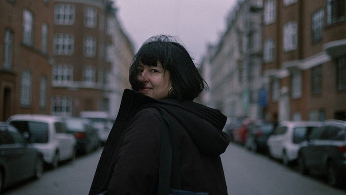 En person i en hættetrøje står og smiler på en svagt oplyst bygade med parkerede biler og bygninger.