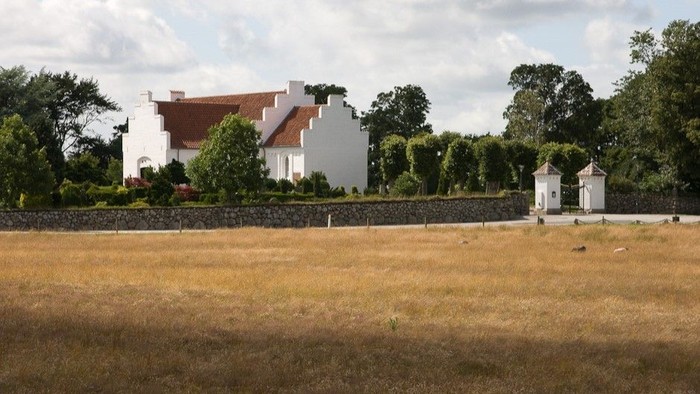 En stor, hvidkalket landhus med røde tag og en mur omgivet af en grøn skov.