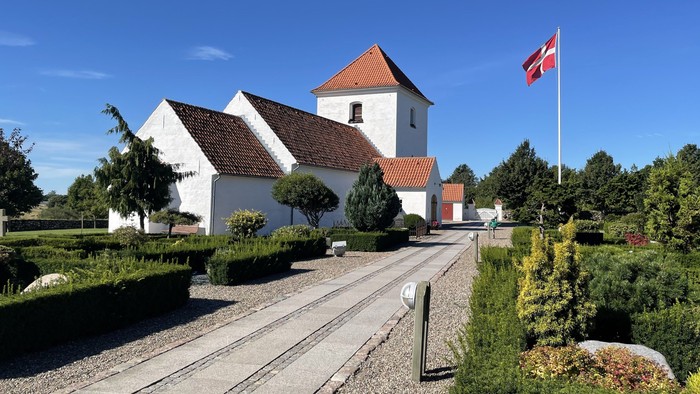 En hyggelig hvid kirke med rødt tag og en flagstang med et nationalflag, omgivet af velplejet grønt.