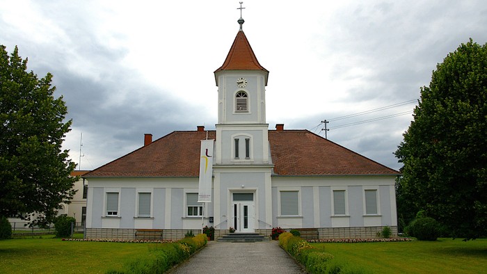 Weißes Kirchengebäude mit hohem Turm und roter Ziegeldach, umgeben von grünem Rasen und Bäumen