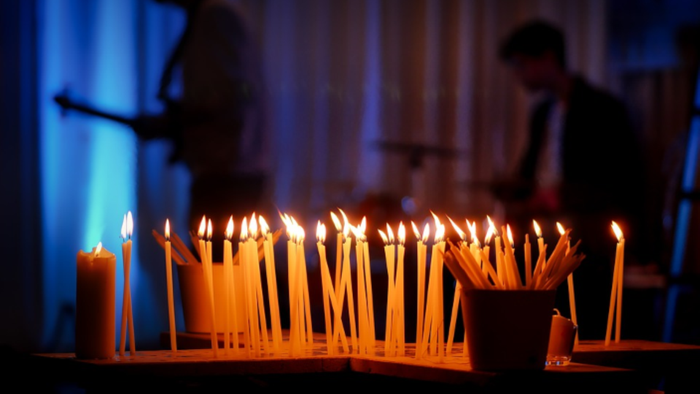 Viele brennende Kerzen auf einem Tisch, zwei Personen im Hintergrund.
