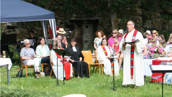 Freiluftzeremonie mit einem Priester, der zu einem aufmerksamen Publikum spricht, das auf Gartenstühlen sitzt.
