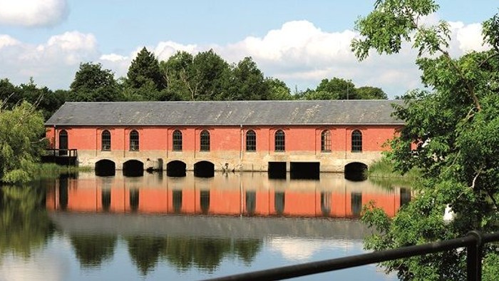 Historisk rødstensbygning med buede åbninger, der spejler sig i stille vand, omgivet af frodig grønne omgivelser.