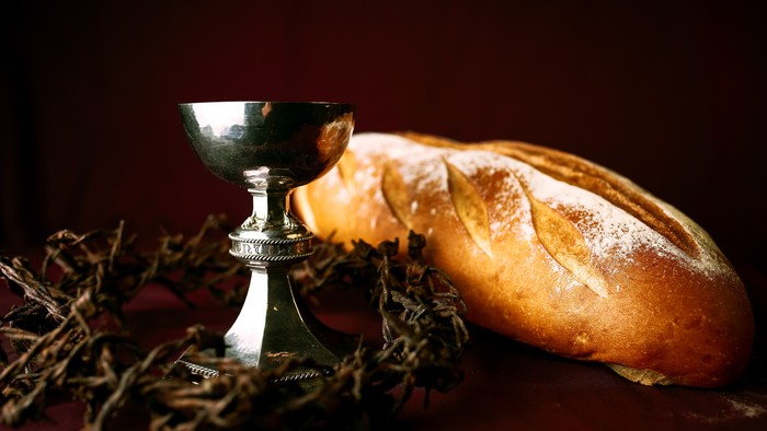 A silver chalice and a loaf of bread are displayed with herbs.