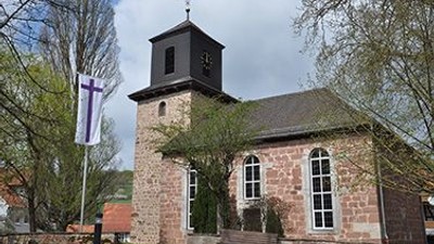 Steinerne Kirche mit hohem Turm und Uhr, umgeben von Bäumen und Gras.
