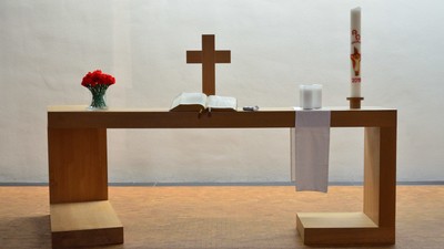 Minimalistischer Altar mit Kreuz, Blumen, Kerze und offenem Buch auf einem hölzernen Lesepult.