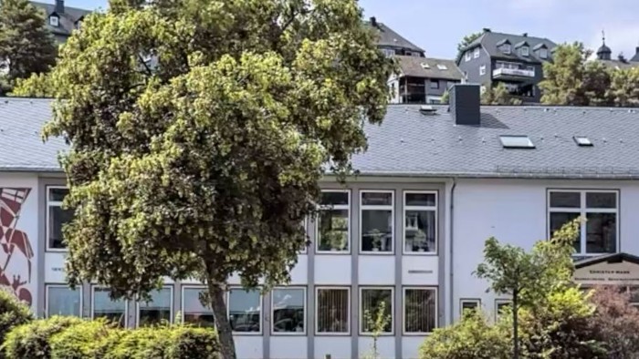 Ein modernes Gebäude mit großen Fenstern und einem Baum davor, umgeben von anderen Häusern auf einem Hügel.