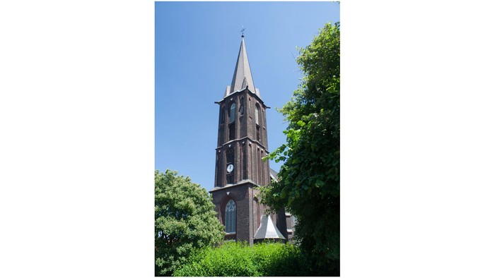 Ein hoher Kirchturm mit Uhr und spitzem Dach vor grünen Bäumen und blauem Himmel