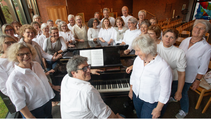 Eine fröhliche Chorgruppe, die sich gemeinsam mit ihrem Dirigenten an einem Klavier in einem Proberaum fotografieren lässt.
