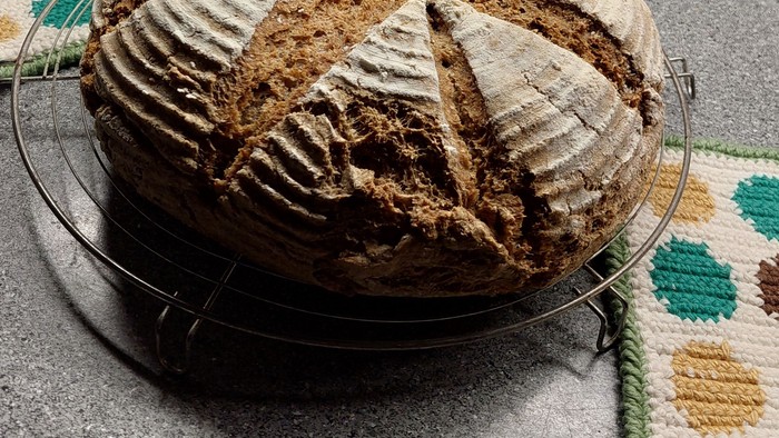 Frisch gebackenes Sauerteigbrot mit Kreuzschnitten auf einem Kühlgitter neben einer Backschüssel und einem Ofen.