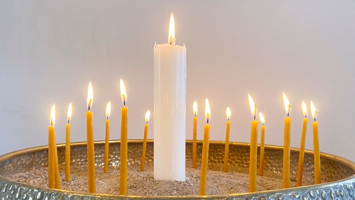 En høj hvid stearinlys står centreret blandt otte mindre tændte lys i en dekorativ metalholder.