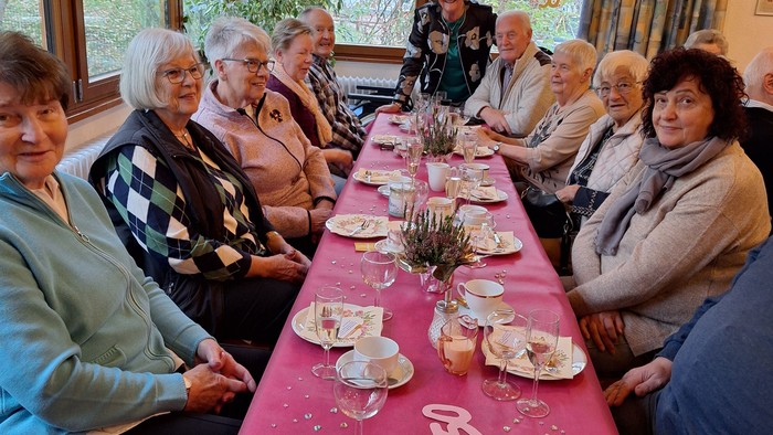 Gruppe älterer Menschen, die bei einer Teeparty mit verzierten Tellern und Tassen an einem langen Tisch genießen.