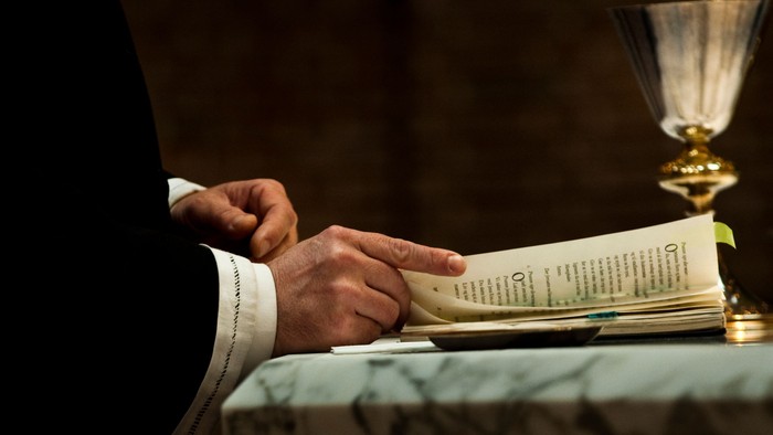 En person i religiøst tøj læser fra en åben bog, med en kalk stående i nærheden på en marmoroverflade.