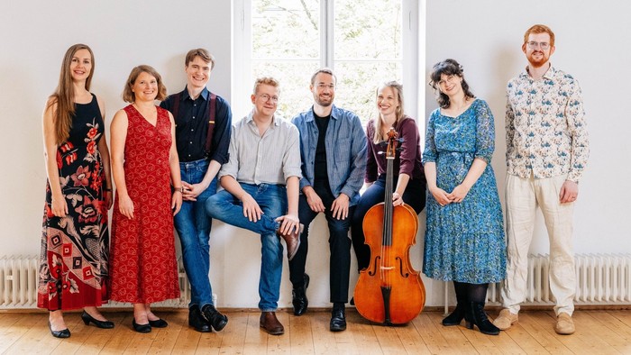 Eine vielfältige Gruppe von sieben Musiker:innen, die sich gemeinsam mit einem Cello zwischen ihnen drinnen aufstellen.