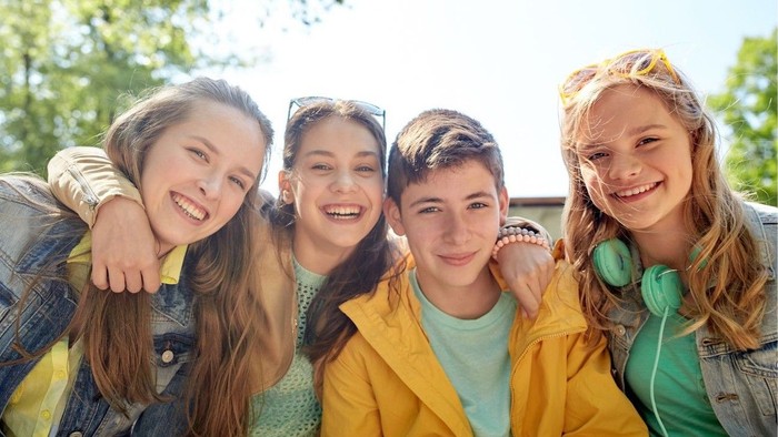 Fire unge venner smiler til kameraet, tre piger og en dreng, ude ved solskinnet
