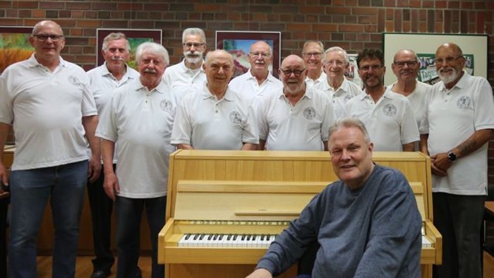 Ein Mann sitzt an einem Klavier, umgeben von einer Gruppe älterer Männer in weißen Hemden