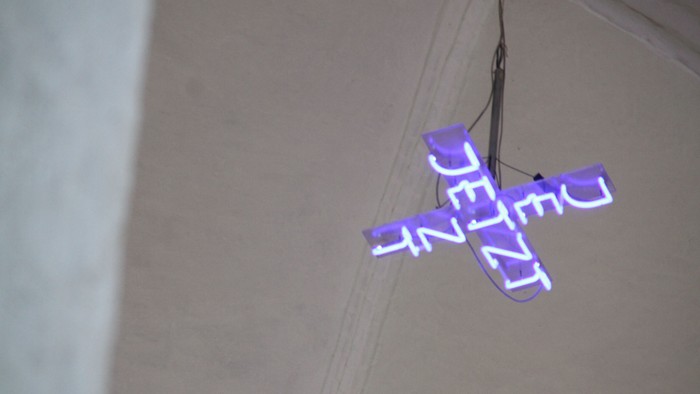 Eine leuchtende Neon-Kreuz mit dem Wort „JESUS“, das in lila und weißen Buchstaben erstrahlt.