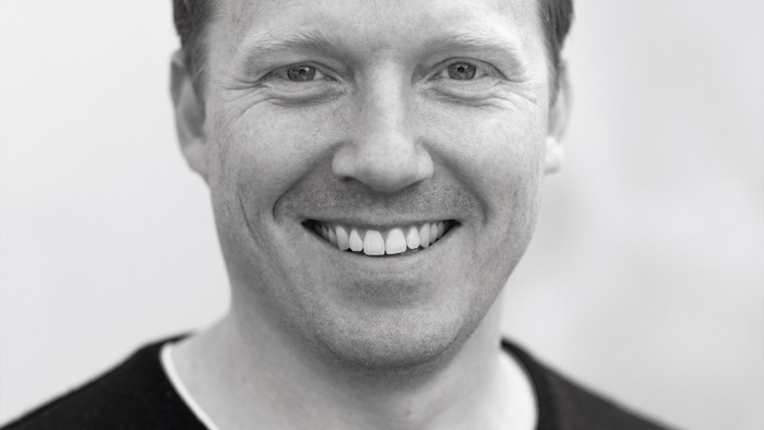 Smilende portræt af en mand i sort skjorte mod en ensfarvet baggrund.