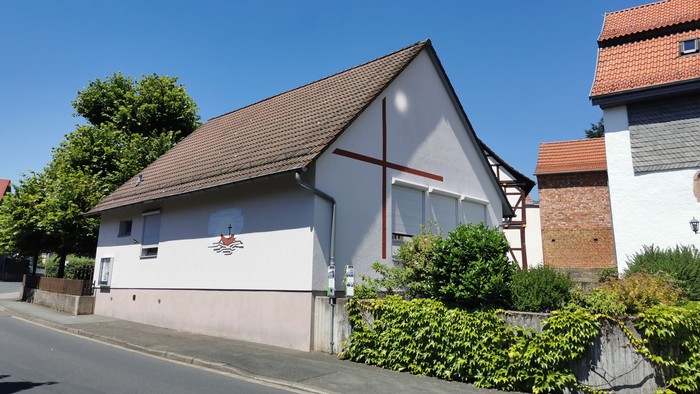 Kleine Kirche mit Kreuz an der Seite, umgeben von Büschen und Bäumen, neben einem Ziegelgebäude.
