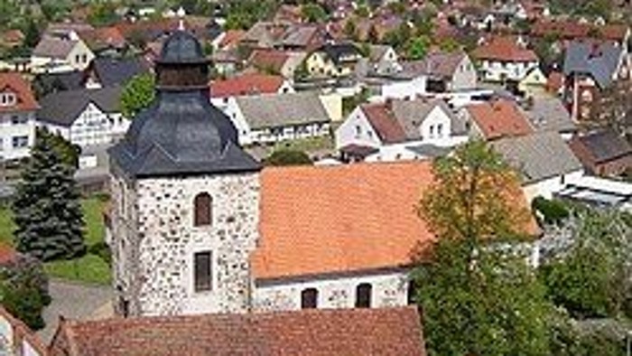 Luftaufnahme eines malerischen Dorfes mit historischer Steinkirche und rotgedeckten Häusern, umgeben von Grünflächen.