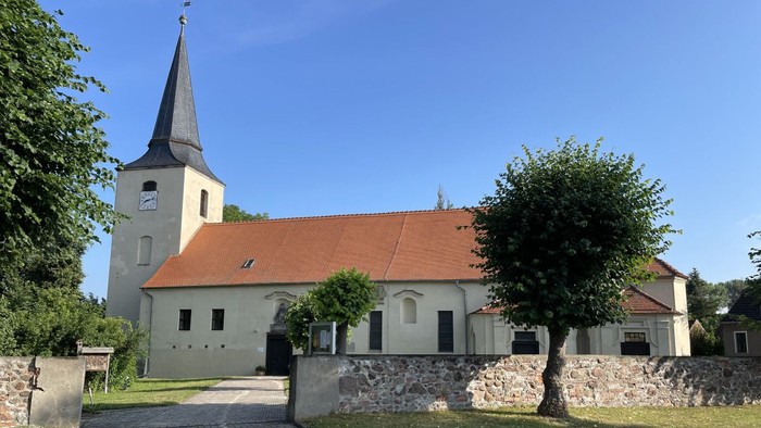 Idyllische Kirche mit hohem Kirchturm und rotem Ziegeldach, umgeben von üppigem Grün und einem Kopfsteinpflasterweg.