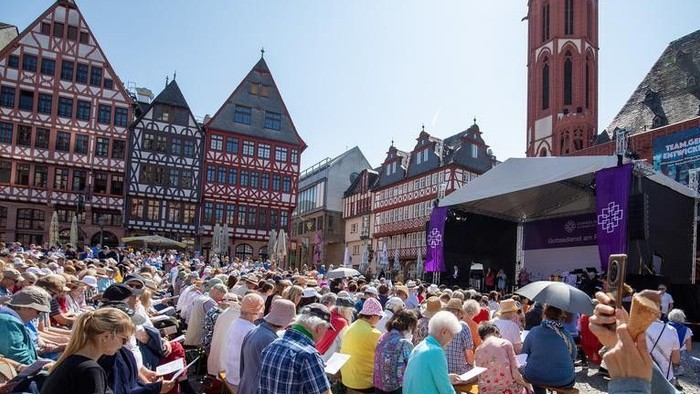 Eine lebhafte Outdoor-Veranstaltung mit einer vielfältigen Menschenmenge, die sitzt und steht und eine Bühnenaufführung auf einem historischen Stadtplatz verfolgt.