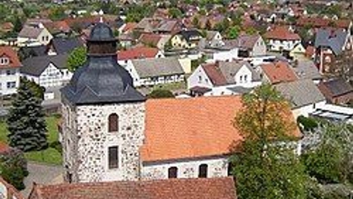 Luftaufnahme eines malerischen Dorfes mit historischer Steinkirche und rotgedeckten Häusern, umgeben von Grünflächen.