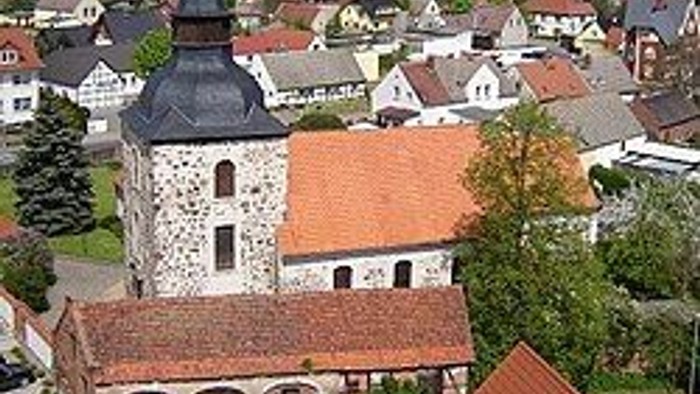 Luftaufnahme eines malerischen Dorfes mit historischer Steinkirche und rotgedeckten Häusern, umgeben von Grünflächen.