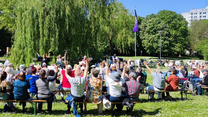 Ein lebhaftes Treffen im Freien mit Menschen, die auf Bänken sitzen und unter Bäumen singen oder jubeln.