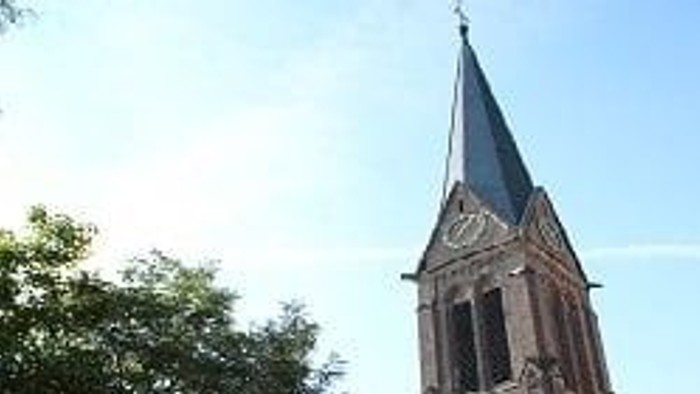 Ein hoher Backsteinkirchturm mit Uhr und Spitze, der sich über grün belaubte Bäume erhebt.